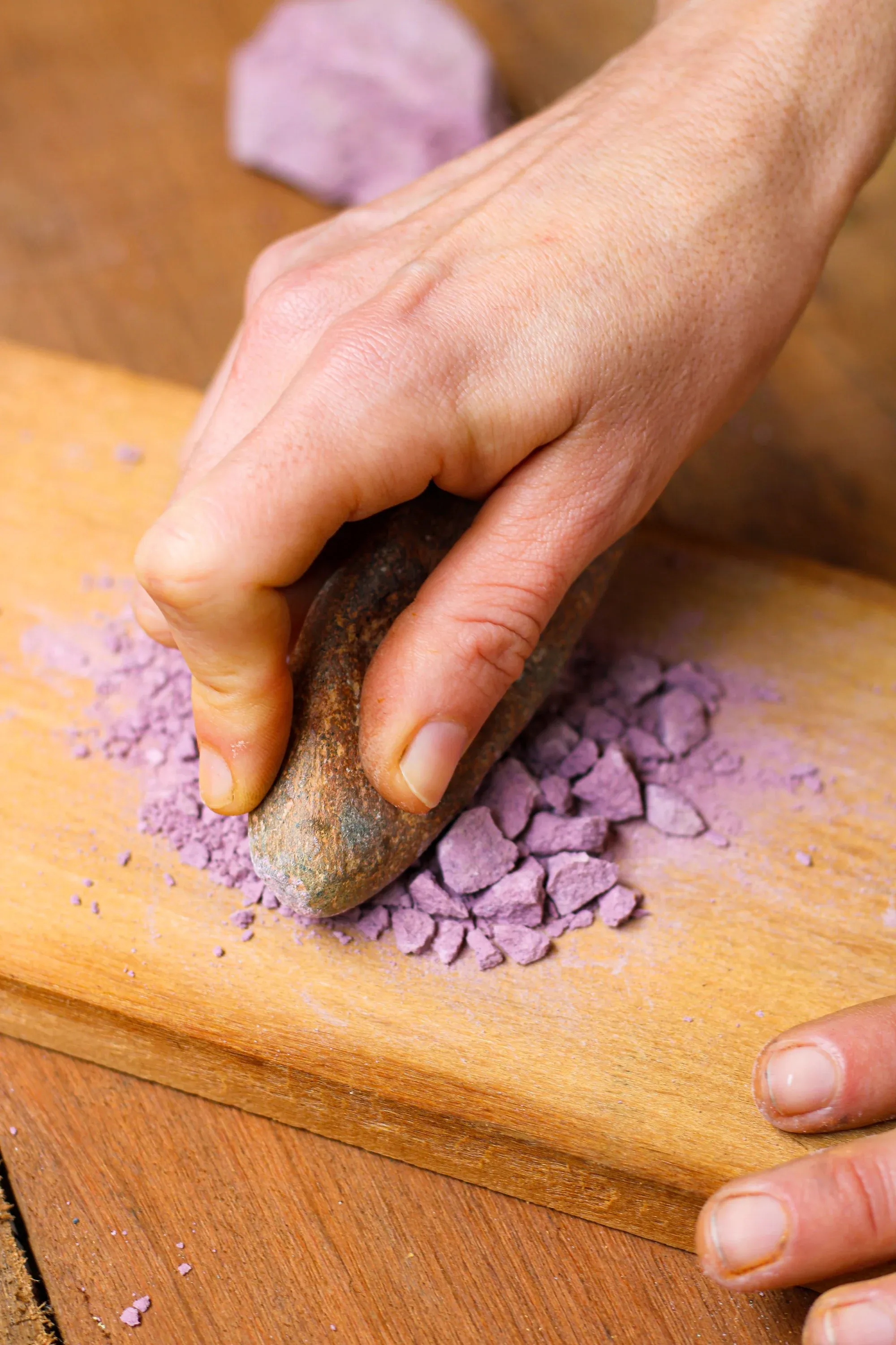 Mão macerando pigmento violeta com pilão de pedra sobre tábua de madeira