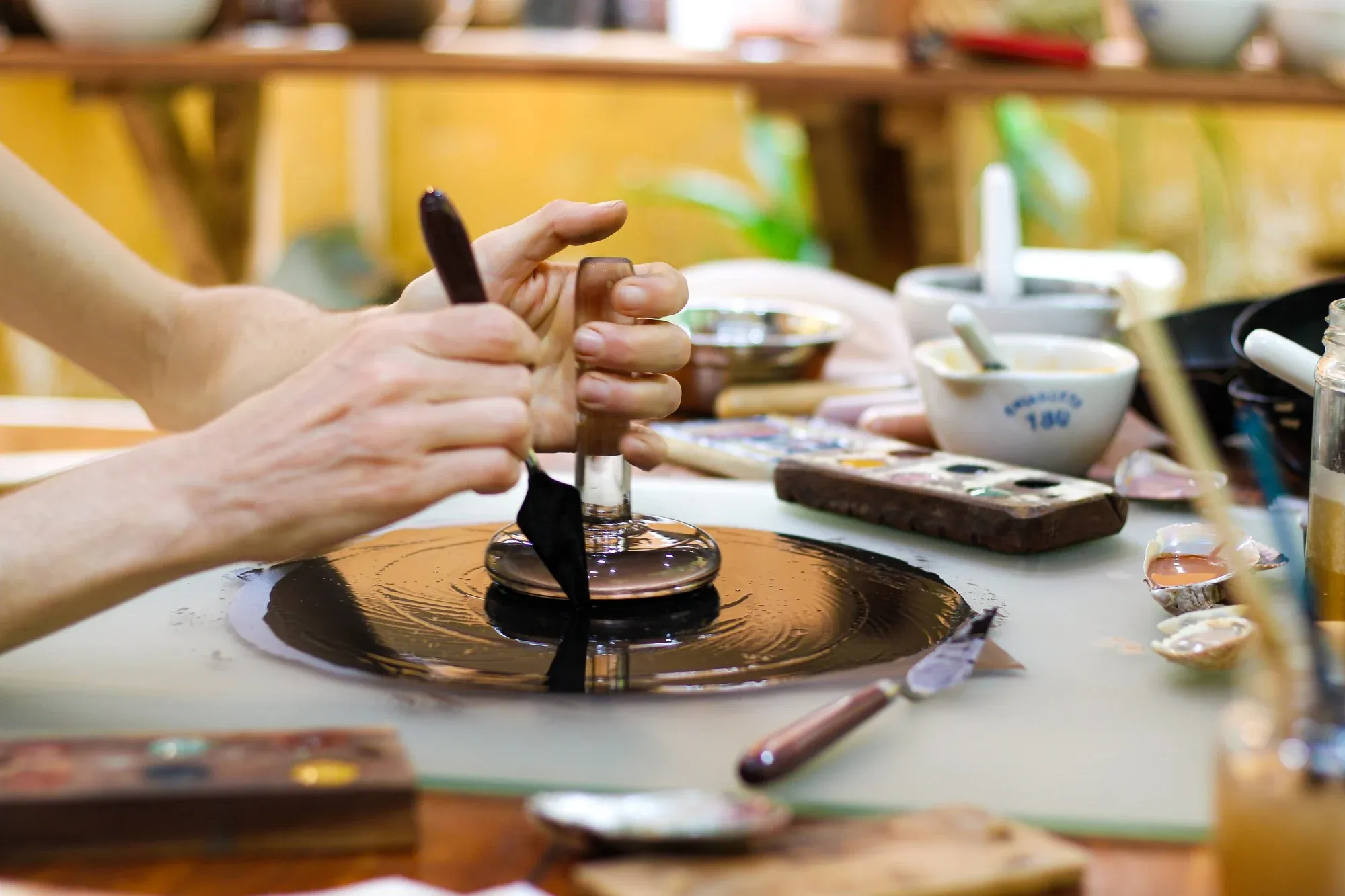 Mãos preparando tinta natural sobre placa de vidro no ateliê, com pigmentos e espátulas ao redor